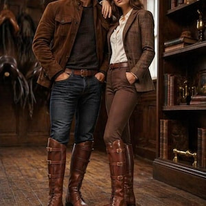 May include: A couple in a room with a wooden floor, wearing brown leather riding boots. The man wears a brown suede jacket, dark jeans, and a dark shirt. The woman wears a brown tweed blazer, brown trousers, and a white shirt. Bookshelves are in the background.