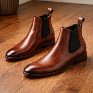 Puede incluir: Un par de botines Chelsea de cuero marr&oacute;n pulido. Los botines tienen un dise&ntilde;o elegante con paneles laterales el&aacute;sticos y una leng&uuml;eta en la parte trasera. Est&aacute;n sobre un suelo de madera.