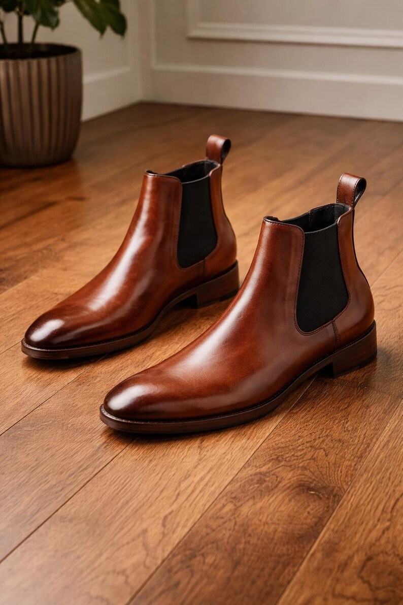 Puede incluir: Un par de botines Chelsea de cuero marr&oacute;n pulido. Los botines tienen un dise&ntilde;o elegante con paneles laterales el&aacute;sticos y una leng&uuml;eta en la parte trasera. Est&aacute;n sobre un suelo de madera.