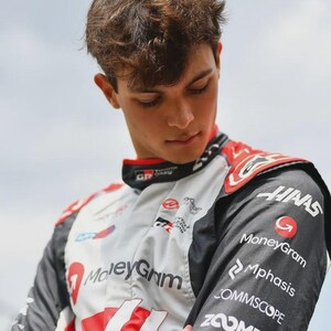 Puede incluir: Un traje de carreras en blanco, negro y rojo, con logotipos de patrocinadores como MoneyGram y Haas. El traje tiene un cuello alto y lo lleva una persona con cabello casta&ntilde;o, mirando hacia abajo. El logotipo de Toyota GR es visible.