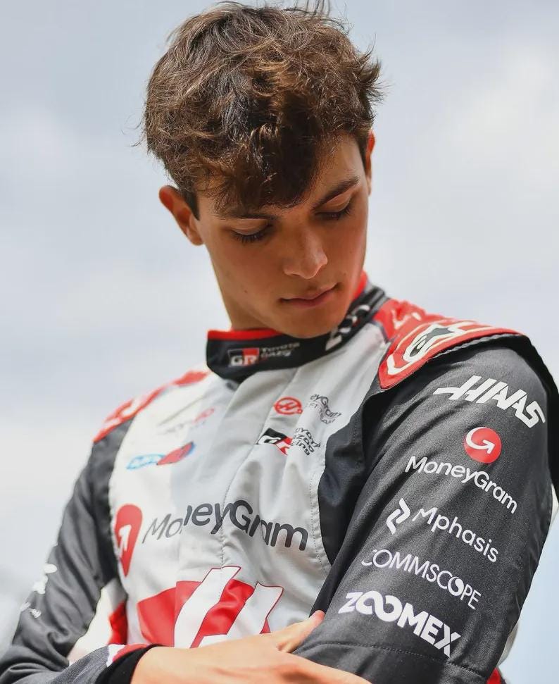 Puede incluir: Un traje de carreras en blanco, negro y rojo, con logotipos de patrocinadores como MoneyGram y Haas. El traje tiene un cuello alto y lo lleva una persona con cabello casta&ntilde;o, mirando hacia abajo. El logotipo de Toyota GR es visible.