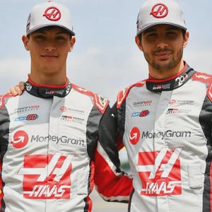 Puede incluir: Dos personas con trajes de carreras y gorras a juego. Los trajes son principalmente blancos con detalles rojos y negros, con logotipos de patrocinadores como "MoneyGram" y "HAAS". Las gorras son grises con un logotipo rojo. Est&aacute;n de pie al aire libre en un d&iacute;a soleado.