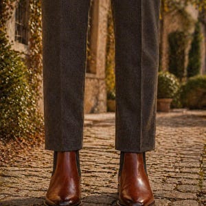 Puede incluir: Botas Chelsea de cuero marr&oacute;n con pantalones grises oscuros. Las botas tienen un acabado pulido y est&aacute;n sobre un camino adoquinado. Los pantalones son de color gris oscuro.