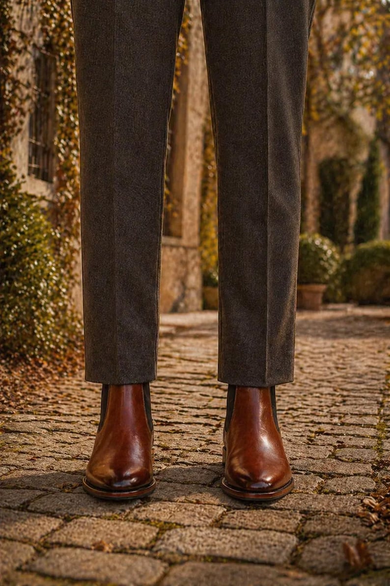 Puede incluir: Botas Chelsea de cuero marr&oacute;n con pantalones grises oscuros. Las botas tienen un acabado pulido y est&aacute;n sobre un camino adoquinado. Los pantalones son de color gris oscuro.