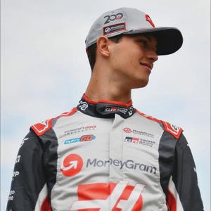 Puede incluir: Gorra de b&eacute;isbol gris claro con visera negra y detalles rojos, con el n&uacute;mero "200" y el logotipo de "MoneyGram". La persona lleva un traje de carreras con los logotipos de "MoneyGram" y "HAAS", y detalles rojos y negros.