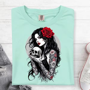 May include: A light mint green t-shirt featuring a graphic illustration of a person with long black hair, a red rose in their hair, and floral tattoos on their arms, holding a human skull. The design includes a circular gray background element.