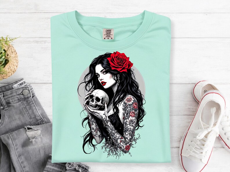 May include: A light mint green t-shirt featuring a graphic illustration of a person with long black hair, a red rose in their hair, and floral tattoos on their arms, holding a human skull. The design includes a circular gray background element.