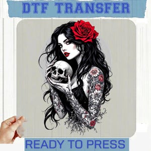 May include: A DTF TRANSFER READY TO PRESS graphic featuring a person with long black hair, a red rose in their hair, and red lipstick, holding a human skull. The person has floral tattoos on their arms and wears a black top against a circular background.