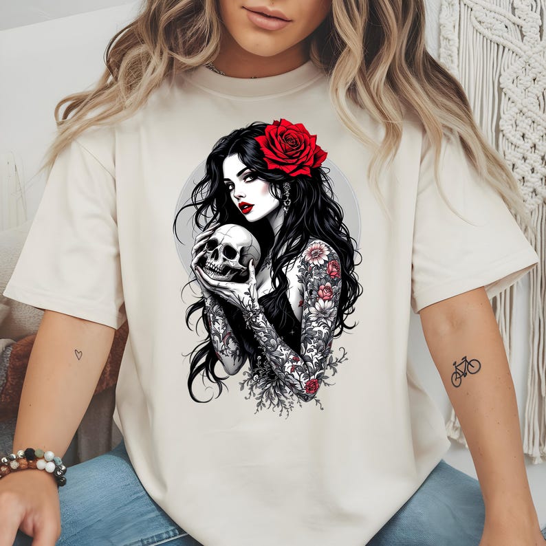 May include: A beige short-sleeve t-shirt featuring a graphic illustration of a person with long black hair, a red rose in their hair, and red lipstick. The person is holding a human skull and has floral tattoos on their arms and shoulders.