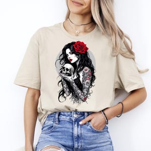 May include: A beige short-sleeve t-shirt featuring a graphic print of a person with long black hair, a red rose in their hair, and floral tattoos on their arms, holding a human skull against a circular gray background.