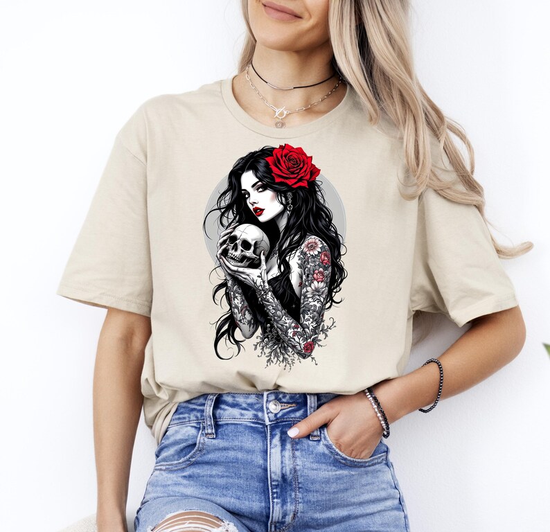May include: A beige short-sleeve t-shirt featuring a graphic print of a person with long black hair, a red rose in their hair, and floral tattoos on their arms, holding a human skull against a circular gray background.