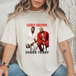 May include: A cream-colored short-sleeve t-shirt featuring a graphic print of Chris Brown and Usher. The text CHRIS BROWN appears in red above the figures, while USHER TERRY is printed in black below. A gold VS emblem sits between the two men.