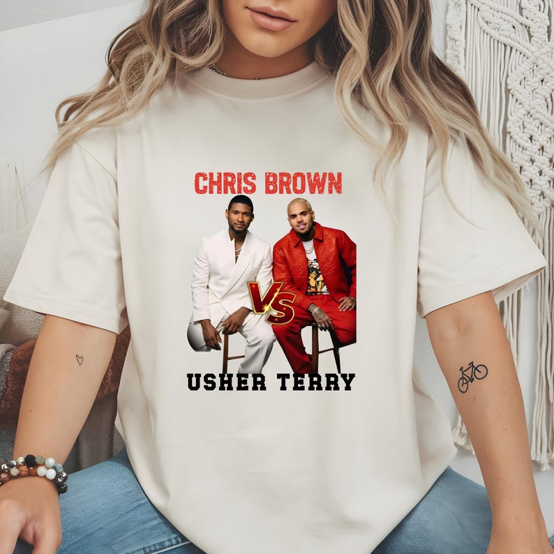 May include: A cream-colored short-sleeve t-shirt featuring a graphic print of Chris Brown and Usher. The text CHRIS BROWN appears in red above the figures, while USHER TERRY is printed in black below. A gold VS emblem sits between the two men.