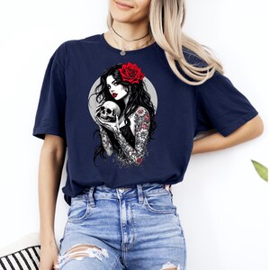 May include: A navy blue t-shirt featuring a graphic print of a person with long dark hair, a red rose in their hair, and red lipstick, holding a skull. The person has detailed floral tattoos on their arms and is set against a light gray circle.