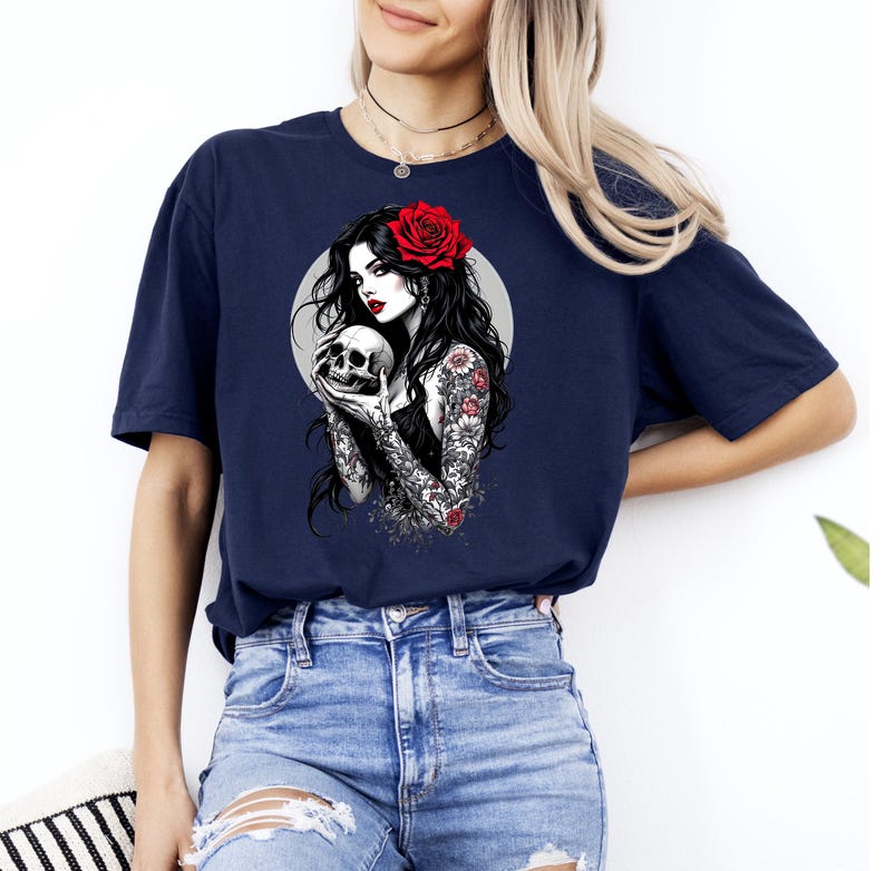 May include: A navy blue t-shirt featuring a graphic print of a person with long dark hair, a red rose in their hair, and red lipstick, holding a skull. The person has detailed floral tattoos on their arms and is set against a light gray circle.