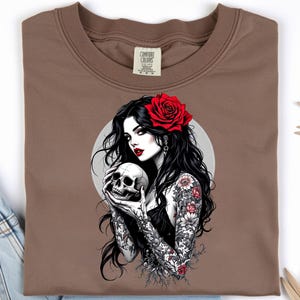 May include: A brown t-shirt featuring a graphic print of a person with long black hair, red lipstick, and a red rose in their hair. The person has floral tattoos on their arms and is holding a human skull. The design includes a circular background.