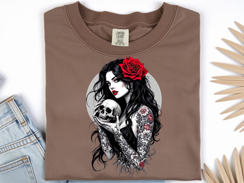 May include: A brown t-shirt featuring a graphic print of a person with long black hair, red lipstick, and a red rose in their hair. The person has floral tattoos on their arms and is holding a human skull. The design includes a circular background.