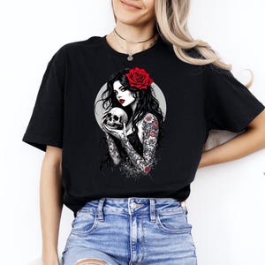 May include: A black t-shirt featuring a graphic print of a person with long dark hair, a red rose in their hair, and red lipstick, holding a skull. The person has detailed floral tattoos on their arms and is set against a circular gray background.