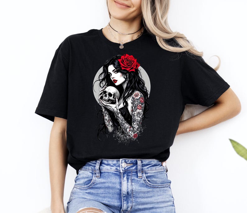 May include: A black t-shirt featuring a graphic print of a person with long dark hair, a red rose in their hair, and red lipstick, holding a skull. The person has detailed floral tattoos on their arms and is set against a circular gray background.