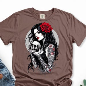 May include: A brown short-sleeve t-shirt featuring a graphic print of a person with long black hair, a red rose in their hair, and red lipstick. The person has floral tattoos on their arms and is holding a human skull in front of a gray circle.
