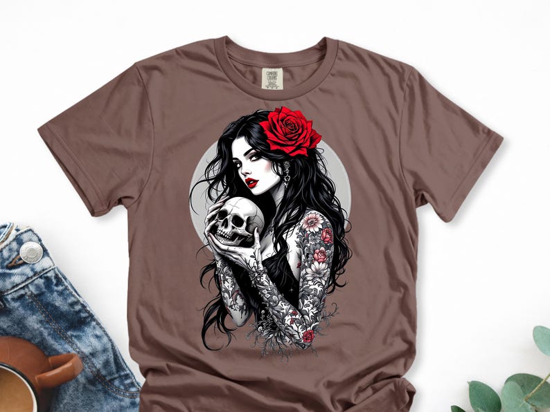 May include: A brown short-sleeve t-shirt featuring a graphic print of a person with long black hair, a red rose in their hair, and red lipstick. The person has floral tattoos on their arms and is holding a human skull in front of a gray circle.