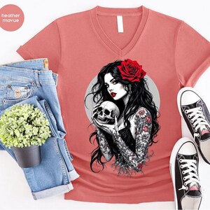 May include: A heather mauve V-neck t-shirt featuring a graphic print of a person with long black hair, a red rose in their hair, and floral tattoos on their arms, holding a human skull. The shirt is displayed with blue denim jeans and sneakers.