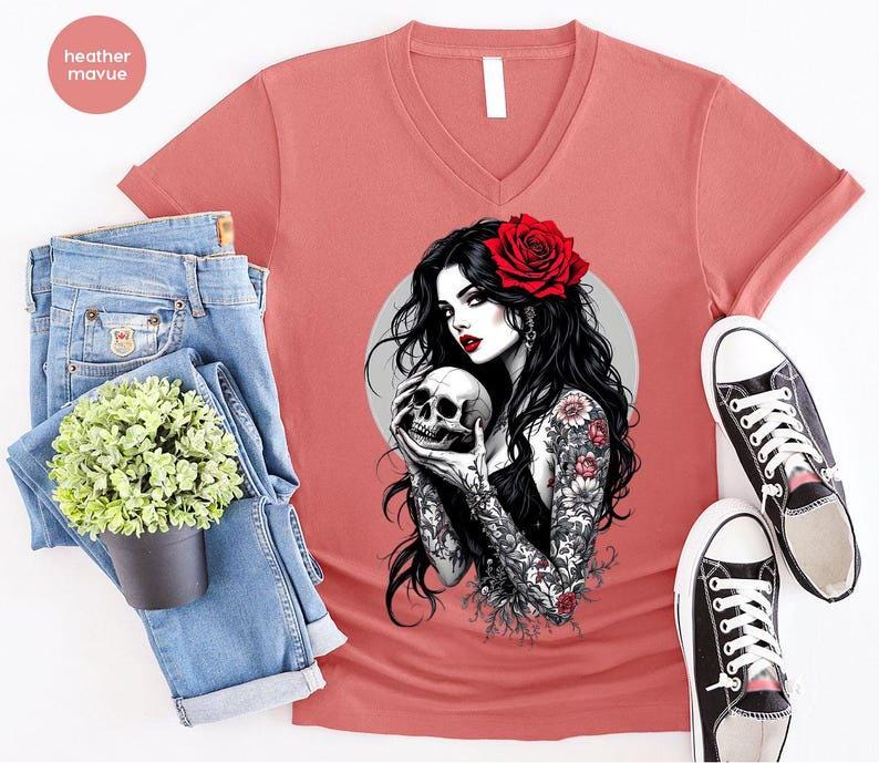 May include: A heather mauve V-neck t-shirt featuring a graphic print of a person with long black hair, a red rose in their hair, and floral tattoos on their arms, holding a human skull. The shirt is displayed with blue denim jeans and sneakers.