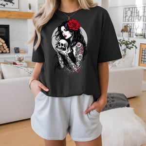 May include: A black short-sleeved t-shirt featuring a graphic print of a person with long dark hair, a red rose in their hair, and red lipstick, holding a skull. The person has floral tattoos on their arms and is set against a light gray circle.