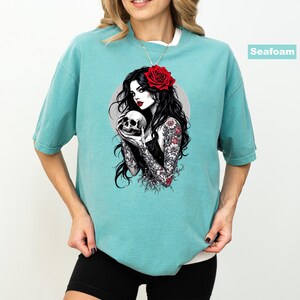 May include: A seafoam green short-sleeve t-shirt featuring a graphic print of a person with long black hair, a red rose in their hair, and floral tattoos on their arms, holding a human skull. The graphic includes a circular gray background element.
