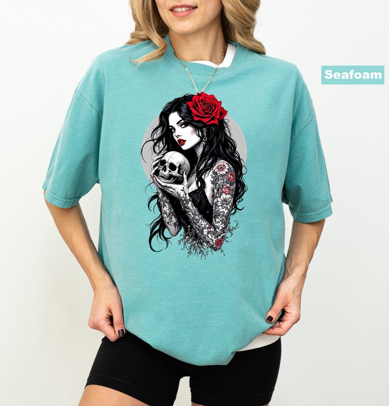 May include: A seafoam green short-sleeve t-shirt featuring a graphic print of a person with long black hair, a red rose in their hair, and floral tattoos on their arms, holding a human skull. The graphic includes a circular gray background element.