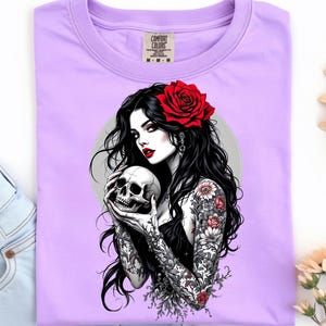 May include: A light purple t-shirt featuring a graphic print of a person with long black hair, a red rose in their hair, and red lipstick, holding a human skull. The person has detailed floral tattoos on their arms and wears a black top.