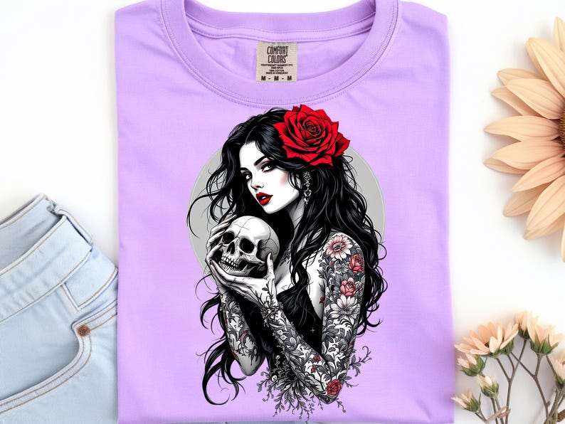 May include: A light purple t-shirt featuring a graphic print of a person with long black hair, a red rose in their hair, and red lipstick, holding a human skull. The person has detailed floral tattoos on their arms and wears a black top.