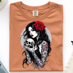 May include: A burnt orange t-shirt featuring a graphic print of a person with long black hair, a red rose in their hair, and intricate floral tattoos on their arms, holding a human skull against a circular gray background.