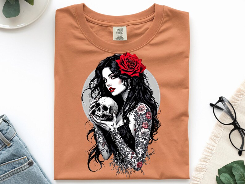 May include: A burnt orange t-shirt featuring a graphic print of a person with long black hair, a red rose in their hair, and intricate floral tattoos on their arms, holding a human skull against a circular gray background.