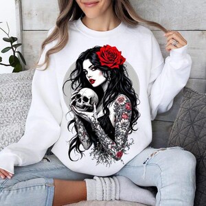 May include: A white crewneck sweatshirt featuring a graphic print of a person with long black hair, a red rose in their hair, and floral tattoos on their arms, holding a human skull. The person has red lips and is set against a circular background.