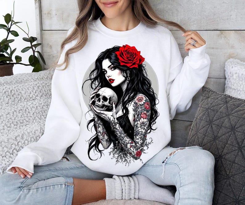 May include: A white crewneck sweatshirt featuring a graphic print of a person with long black hair, a red rose in their hair, and floral tattoos on their arms, holding a human skull. The person has red lips and is set against a circular background.