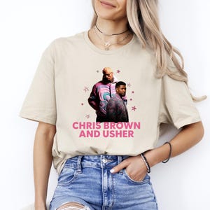 May include: A beige short-sleeved t-shirt featuring a graphic print of Chris Brown and Usher wearing patterned jackets. The design includes small pink star icons and the text CHRIS BROWN AND USHER printed in bold pink capital letters below them.