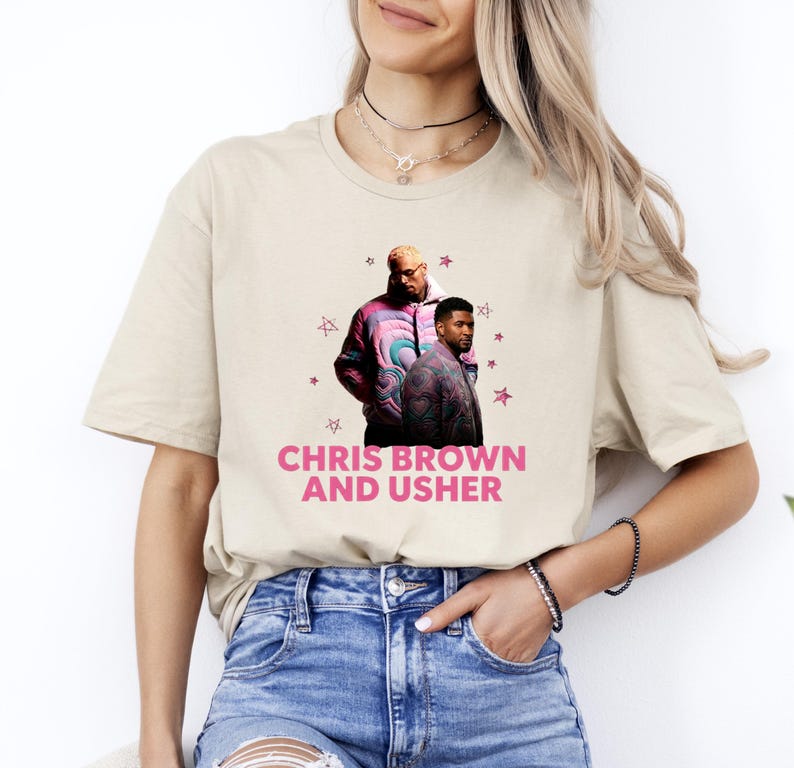 May include: A beige short-sleeved t-shirt featuring a graphic print of Chris Brown and Usher wearing patterned jackets. The design includes small pink star icons and the text CHRIS BROWN AND USHER printed in bold pink capital letters below them.