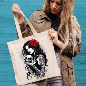 May include: A beige canvas tote bag featuring a graphic illustration of a person with long black hair, a red rose in their hair, and floral tattoos on their arms, holding a human skull. The design includes a circular gray background element.