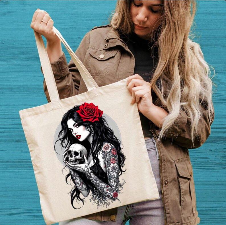 May include: A beige canvas tote bag featuring a graphic illustration of a person with long black hair, a red rose in their hair, and floral tattoos on their arms, holding a human skull. The design includes a circular gray background element.