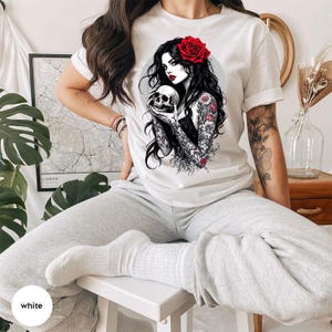 May include: A white short-sleeve t-shirt featuring a graphic illustration of a person with long black hair, a red rose in their hair, and tattoos on their arms, holding a human skull. The person is wearing a black top and has floral tattoos.