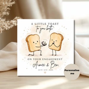 Op de afbeelding: Een vierkante wenskaart met een illustratie van twee sneetjes toast. Eén sneetje knielt en biedt een verlovingsring aan. Tekst: A LITTLE TOAST To you both ON YOUR ENGAGEMENT Aimee & Ben 26TH JUNE 2026. Een rond label bevat de tekst: Personalise me.