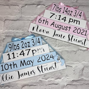 May include: Two wooden signs with blue and pink ribbons, decorated with silver glitter footprints. The top sign reads "7lbs 14oz 3/4 7:14 pm 6th August 2021 Nova Jane Stuart". The bottom sign reads "9lbs 2oz 3/4 11:47pm 10th May 2024 Ollie James Stuart".