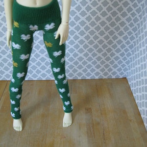 Puede incluir: Leggings verdes con un estampado de tréboles blancos y detalles dorados. Los leggings tienen una cintura ancha y acanalada. La muñeca está de pie sobre una superficie de madera con un fondo gris y blanco estampado.