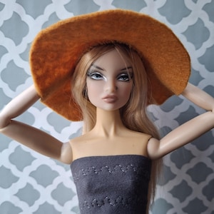 Puede incluir: Una muñeca con el pelo largo y rubio que lleva un sombrero de sol de ala ancha de color naranja quemado y un top gris sin tirantes.