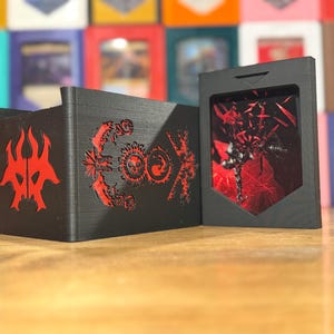 Könnte beinhalten: Schwarzer Kartenhalter und Deckbox mit roten Akzenten. Die Deckbox hat komplizierte Designs und ein Symbol. Der Kartenhalter hat ein Fenster, das ein rotes abstraktes Design zeigt. Im Hintergrund sind weitere Kartenboxen zu sehen.