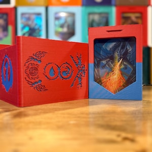 May include: Red and blue card storage boxes. One box features a dragon illustration with flames, while the other has a cut-out design. The boxes are made of a rigid material and are designed to hold playing cards. The background shows other card storage boxes.