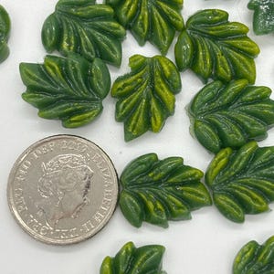 May include: A collection of green leaf-shaped glass embellishments, some with a yellow tint, scattered on a white surface. A British coin is included for scale.