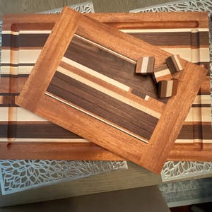 May include: A wooden cutting board with a natural wood finish. The board is made of multiple types of wood, including walnut and maple. The board has a rectangular shape and is approximately 18 inches long and 12 inches wide. The board is sitting on a table with a white tablecloth.