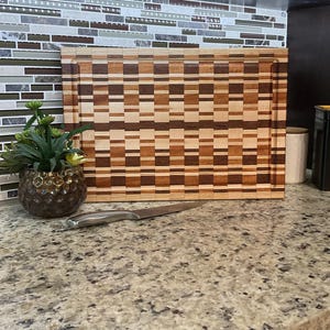 May include: A large wooden cutting board with a checkered pattern in shades of brown and beige. The board is made of different types of wood, creating a unique and rustic look. It is sitting on a granite countertop with a silver knife next to it.